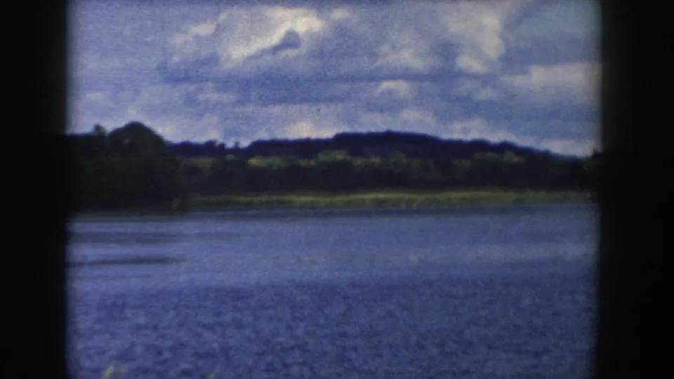 1958 Wisconsin 8mm view across rippled lake water toward a green shoreline and low hills beneath partly sunny clouds.