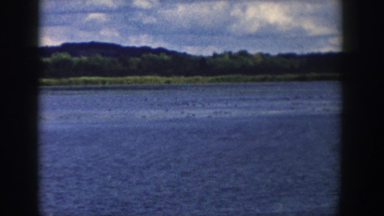 1958 Wisconsin 8mm frame looking across blue lake water to a wooded shoreline and low hills under broken clouds.