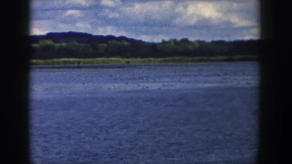 1958 Wisconsin 8mm frame looking across blue lake water to a wooded shoreline and low hills under broken clouds.
