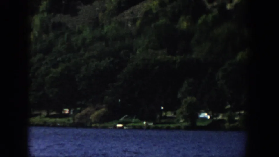 1958 Wisconsin 8mm shoreline view with dense trees, blue water, and small indistinct light objects near the lakeside.