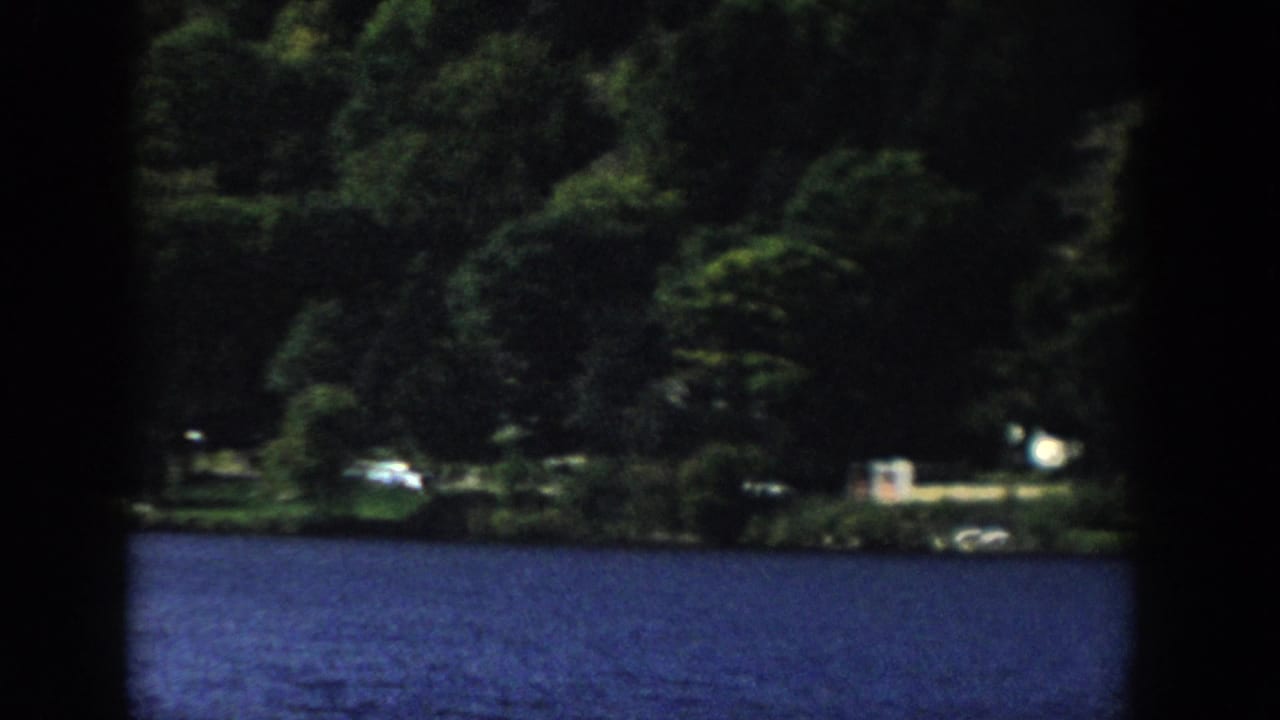 1958 Wisconsin 8mm frame of dark lake water, dense trees, and distant light-toned shapes along the far shore.