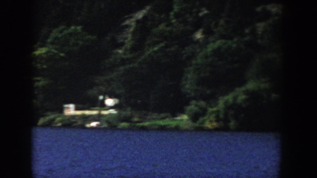 1958 Wisconsin 8mm view across blue lake water toward dense trees and small distant shoreline structures.