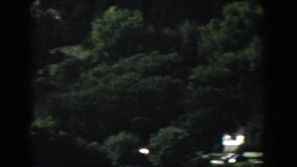 1958 Wisconsin home-movie frame showing a shadowed wooded bank and a small bright vehicle-like shape near the lower edge.