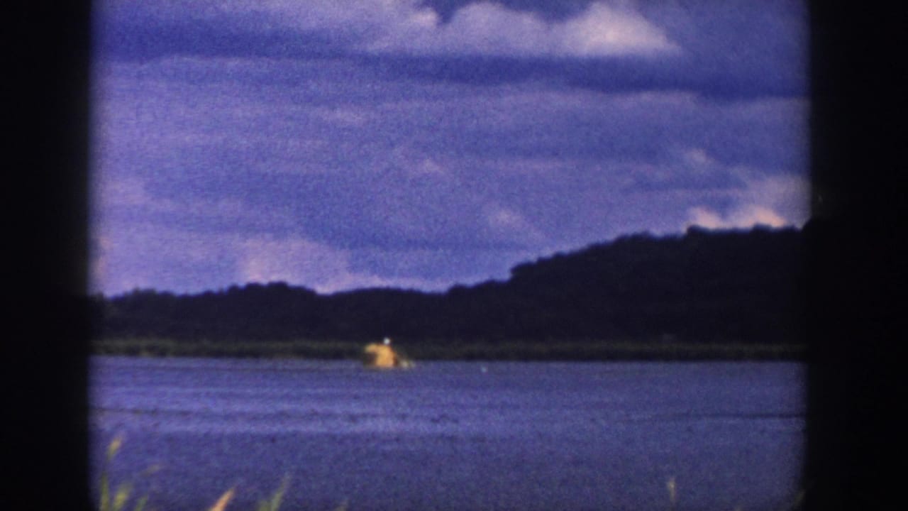 1958 Wisconsin 8mm frame showing blue water, reeds, a distant small boat, wooded hills, and clouded sky through dark film-frame edges.