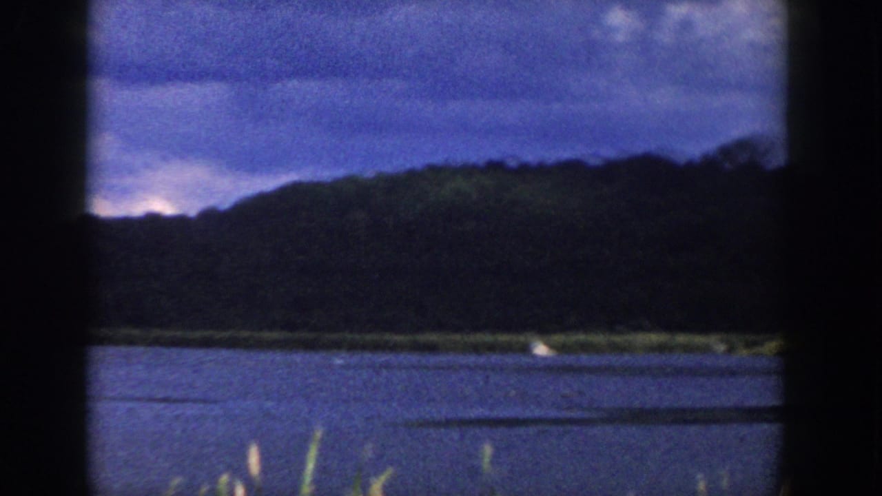 1958 Wisconsin home-movie frame with calm water, a grassy far shore, dark wooded hill, clouded sky, and a small boat at right.