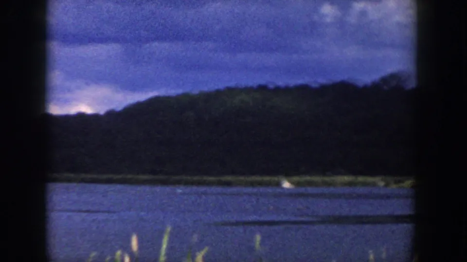 1958 Wisconsin home-movie frame with calm water, a grassy far shore, dark wooded hill, clouded sky, and a small boat at right.