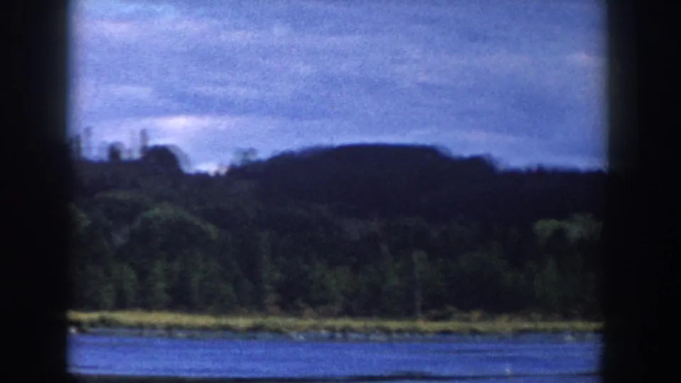1958 Wisconsin home-movie frame of a cloudy lake view, with water, trees, low hills, and dark window framing across the scene.