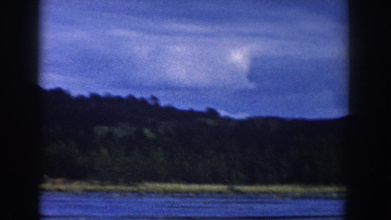 1958 Wisconsin 8mm view of a lake and wooded hillside beneath a cloudy blue sky, framed by dark bars in the foreground.
