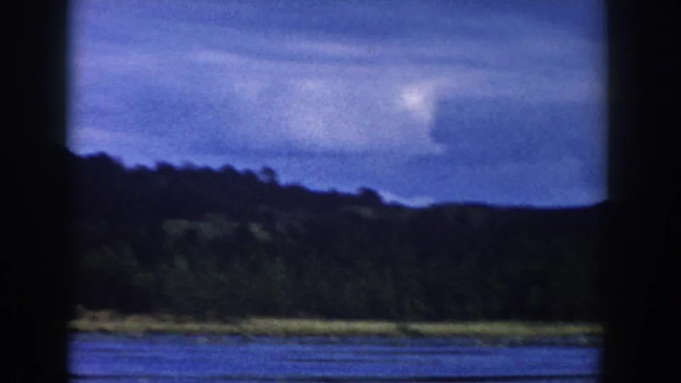 1958 Wisconsin 8mm view of a lake and wooded hillside beneath a cloudy blue sky, framed by dark bars in the foreground.