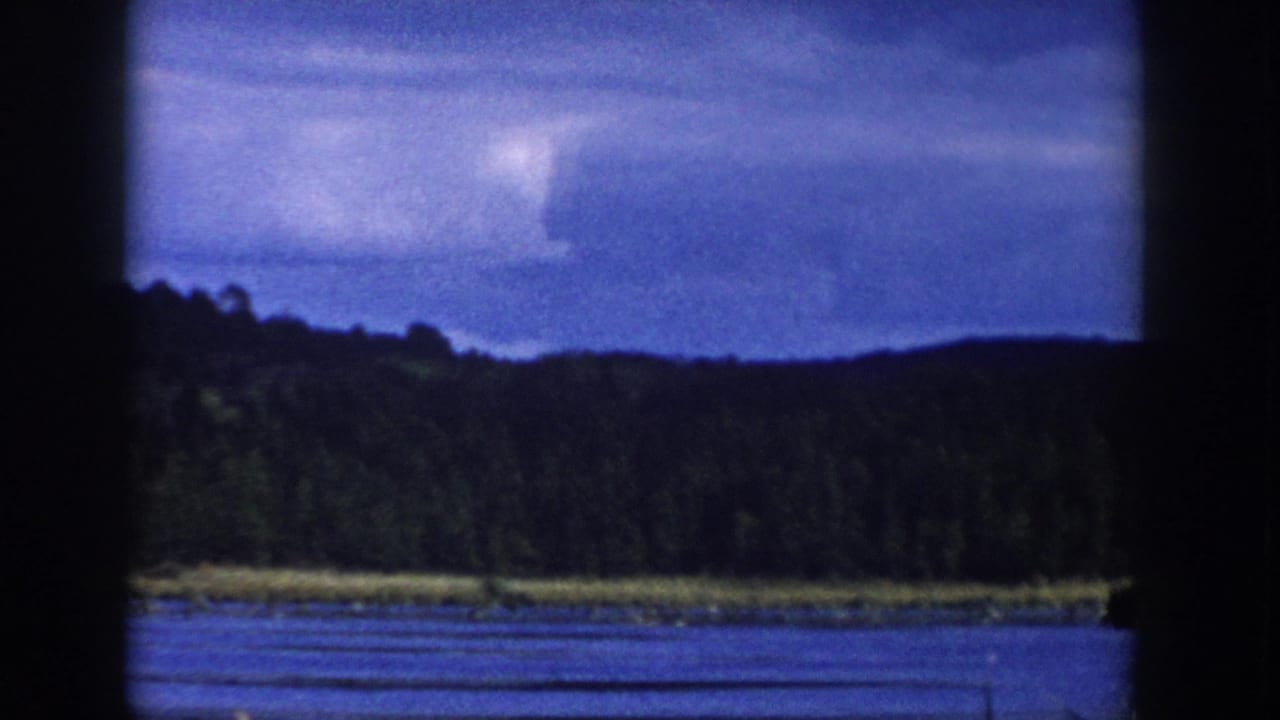 1958 Wisconsin 8mm frame showing a lake, dark tree line, and clouded sky with soft film grain and blue color shift.