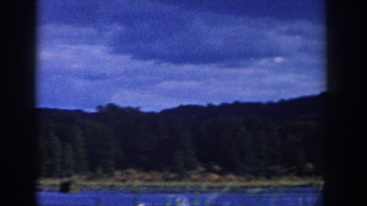 1958 Wisconsin lake scene with forested shoreline, low hills, and a heavy cloud layer seen through soft 8mm exposure.