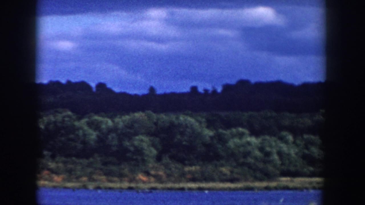 1958 Wisconsin 8mm frame showing a blue lake, leafy shoreline, distant dark ridge, and layered clouds in soft focus.