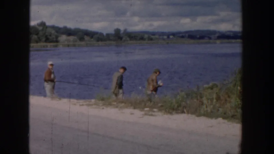 1958 Wisconsin home-movie frame of three boys fishing on a lake bank, with brush, water, trees, and low hills visible.