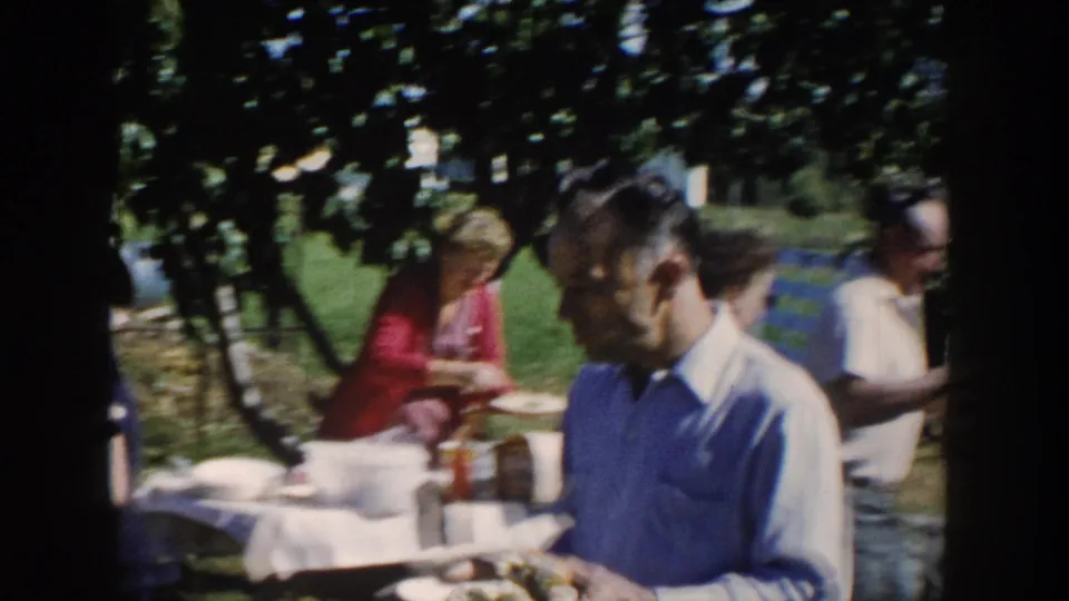 A backyard serving table with adults holding plates during a 1958 Wisconsin family barbecue.
