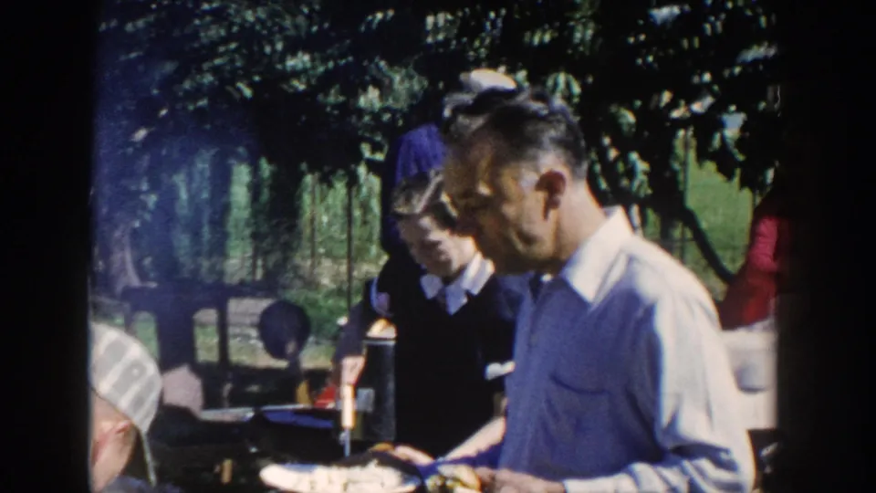 Man in a light shirt near a backyard food table, with other adults partly visible in shade.