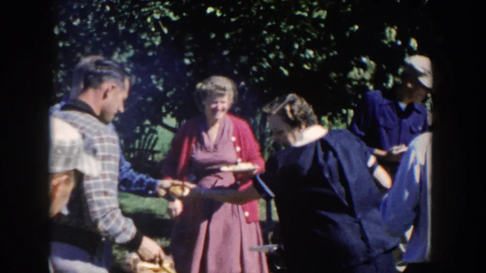 Adults holding plates and serving food in a shaded Wisconsin backyard family barbecue scene.