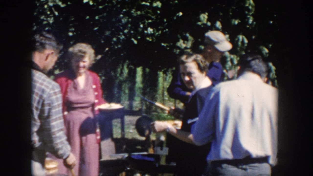 1958 Wisconsin home-movie frame showing older adults with plates around an outdoor barbecue grill in a leafy yard.