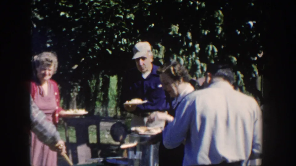 Older adults in 1950s clothing appear to serve food from a small backyard grill in Wisconsin.
