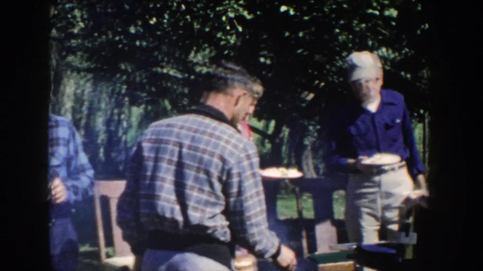 A handheld 8mm frame shows older adults gathered tightly around a backyard barbecue, with trees and lawn behind them.