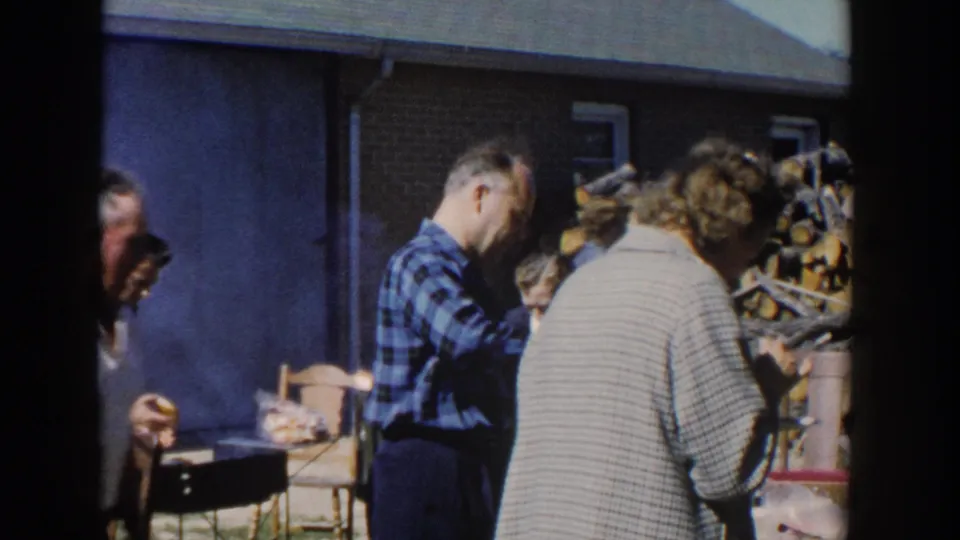 1958 Wisconsin backyard frame with a man in a blue plaid shirt near food, chairs, and other adults beside a house.