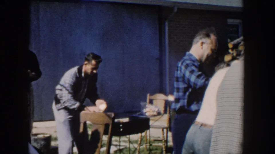 1958 Wisconsin home-movie frame showing a man preparing or serving food as adults stand near chairs and a house wall.