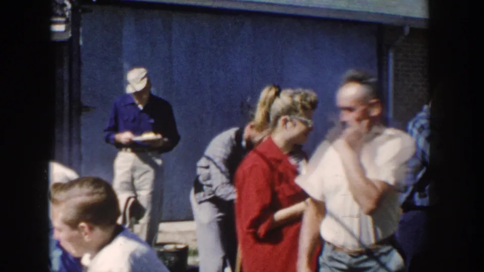 1958 Wisconsin backyard frame showing adults with plates, conversation, a house wall, and a stacked woodpile behind them.