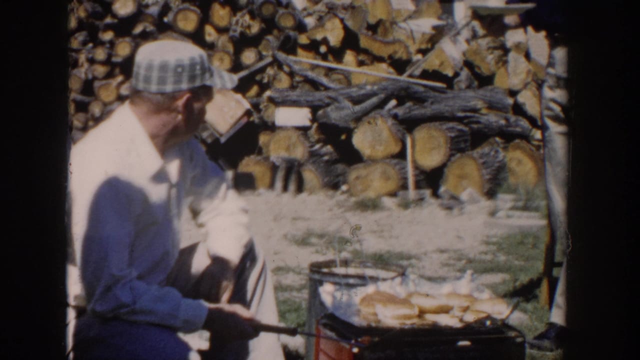 In 1958 Wisconsin home-movie footage, a man in a light shirt and plaid cap tends a small outdoor grill beside stacked firewood.