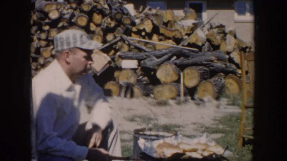 The man sits in profile beside the grill, with buns or food cooking and a large stack of chopped firewood behind him.