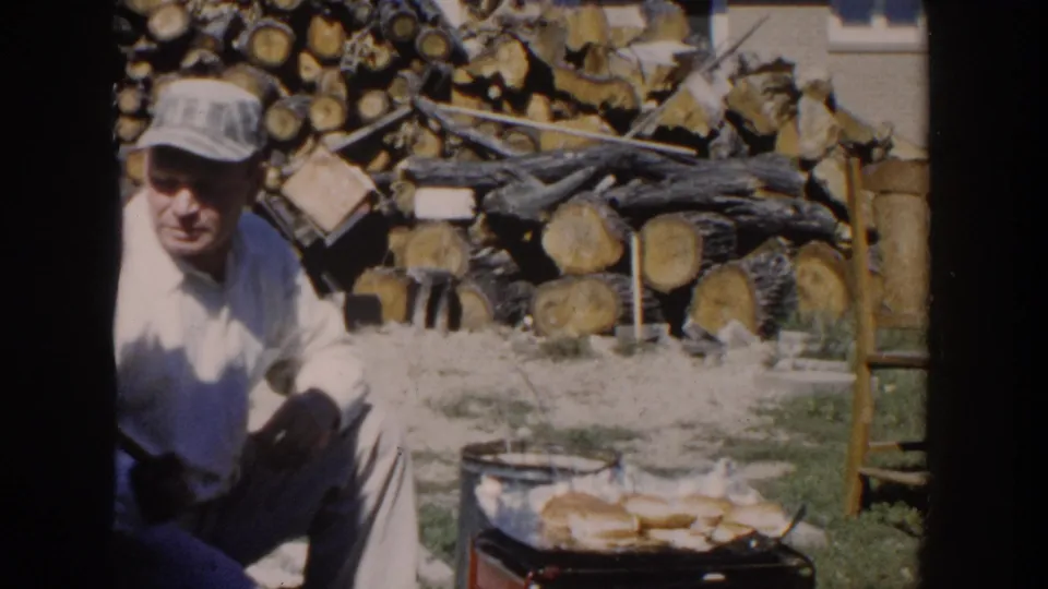 The seated man turns partly toward the camera while the outdoor grill and stacked firewood remain visible in the yard.
