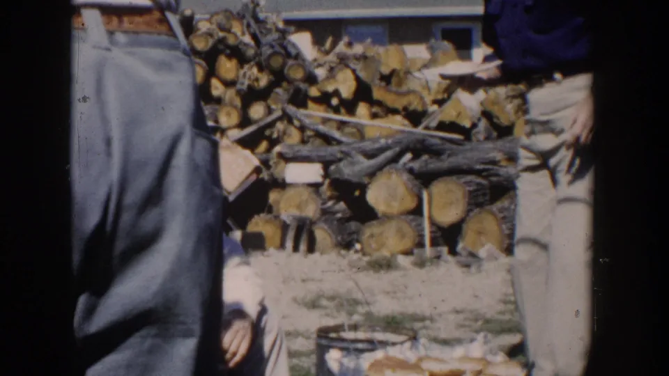A cropped figure stands near the grill while the seated man is partly visible, with stacked firewood filling much of the background.