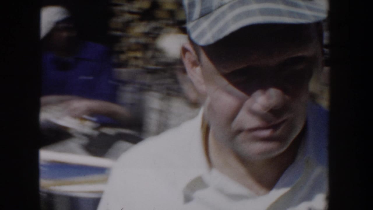 1958 Wisconsin home-movie frame showing a close, shadowed view of an adult man in a light cap and pale collared shirt outdoors.