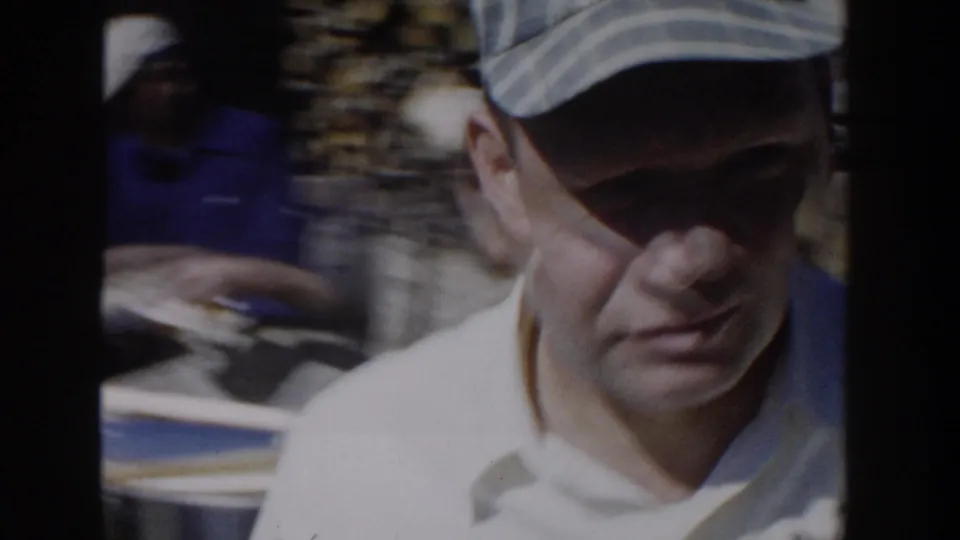 1958 Wisconsin home-movie frame showing a close, shadowed view of an adult man in a light cap and pale collared shirt outdoors.