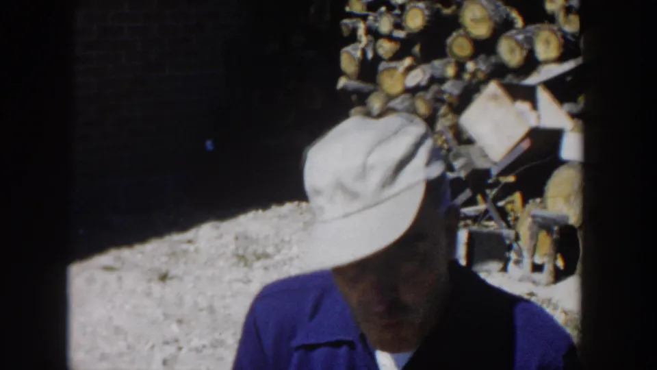 1958 Wisconsin frame of the man looking down in partial shadow, with chopped logs and yard materials behind him.