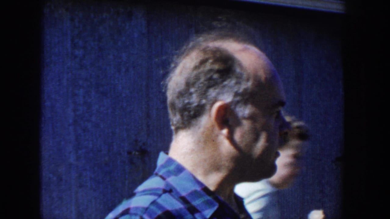 Man in a blue plaid shirt turns in profile with another person partly visible behind him.