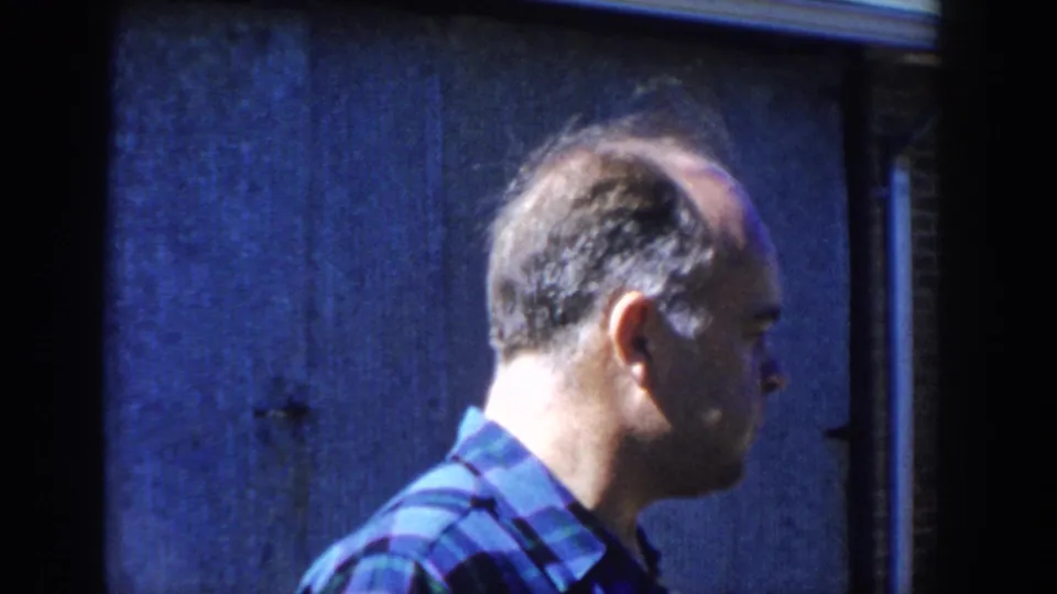 The same adult man in a blue plaid shirt appears lower in the handheld frame as he moves or turns beside a dark blue wooden wall.