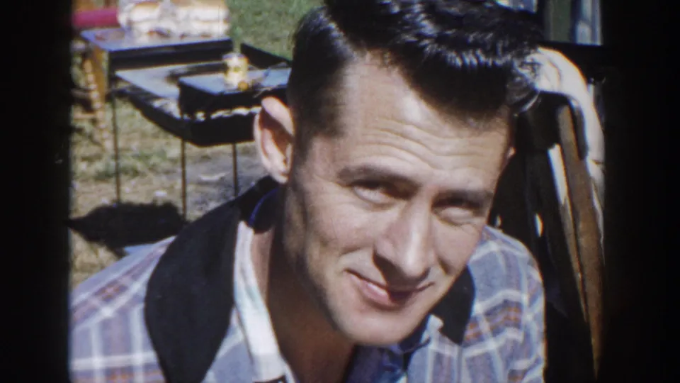 An adult man in a light plaid shirt with a dark collar looks toward the camera in outdoor 1958 Wisconsin home-movie footage, with furniture partly visible behind him.