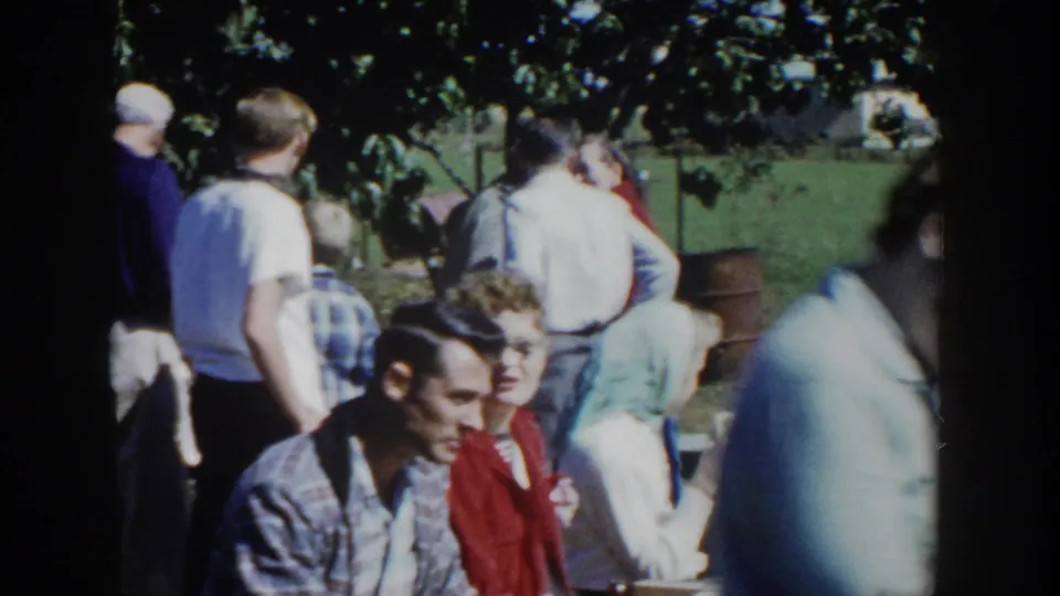Wider 1958 Wisconsin picnic view with seated guests, standing figures, grass, and tree shade in a casual home-movie scene.