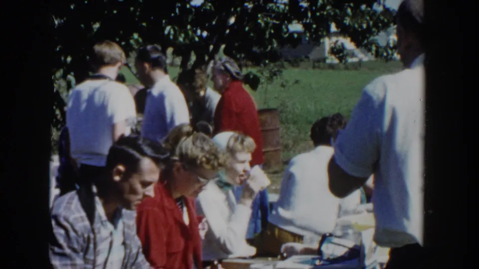 Outdoor picnic frame showing several people seated at tables with others standing nearby in a shaded Wisconsin home-movie scene.