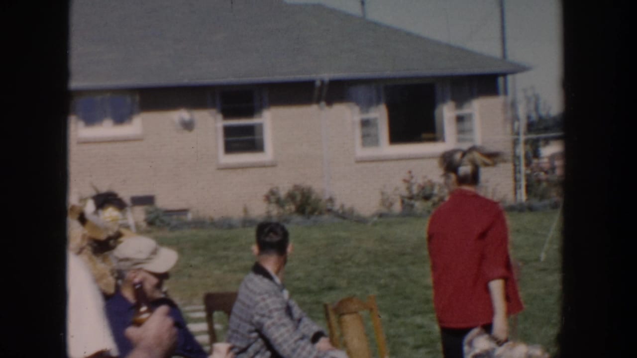 1958 Wisconsin home-movie frame showing adults gathered in a suburban backyard near a brick house, with lawn chairs and people looking offscreen.