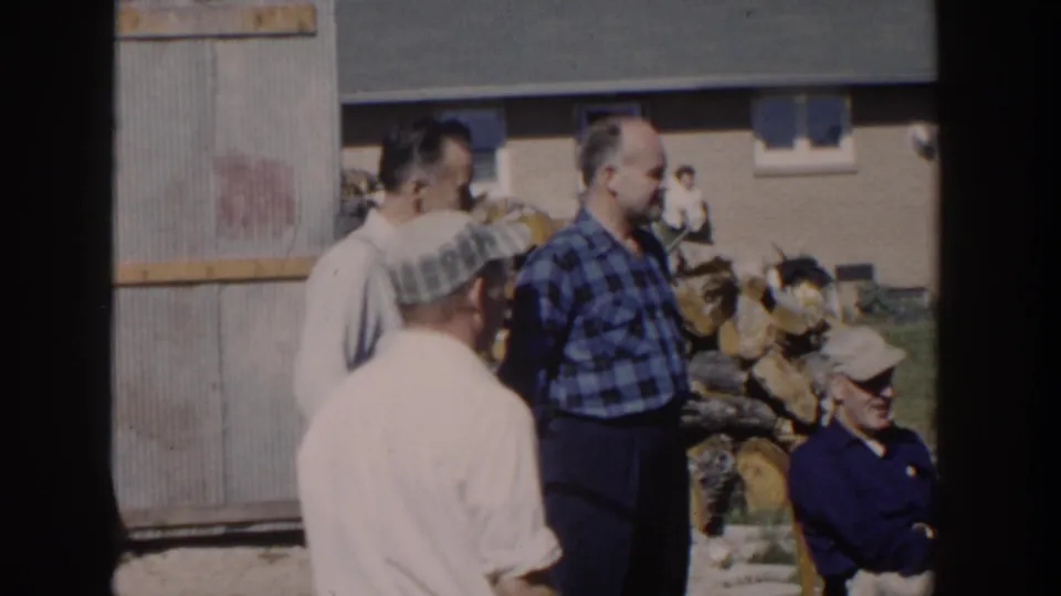 1958 Wisconsin backyard frame with adult men in plaid shirts and caps near a woodpile, shed wall, and brick house background.