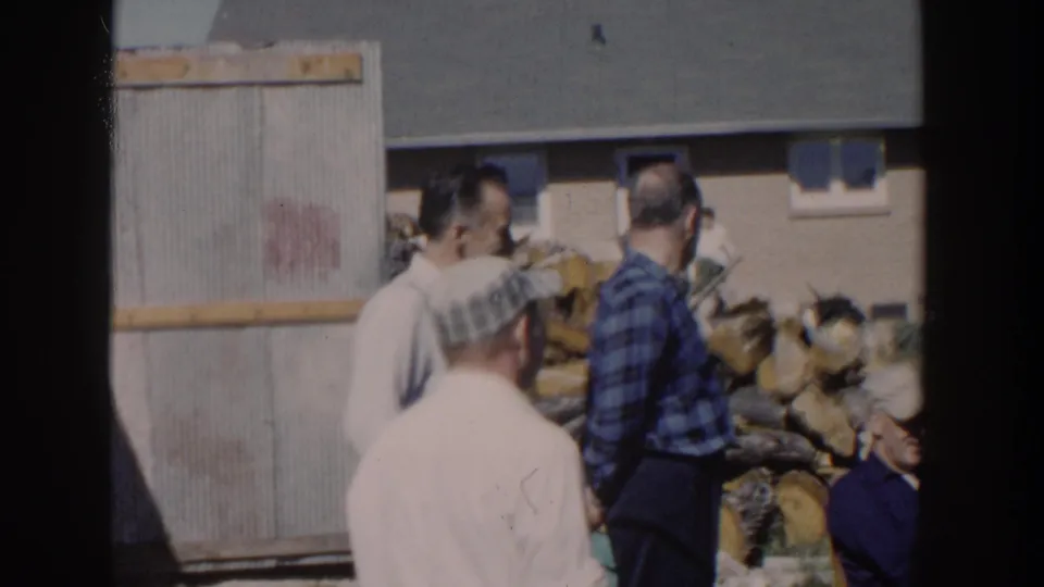 1958 Wisconsin home-movie frame showing adult men gathered near a shed or fence panel, stacked firewood, and a suburban house.