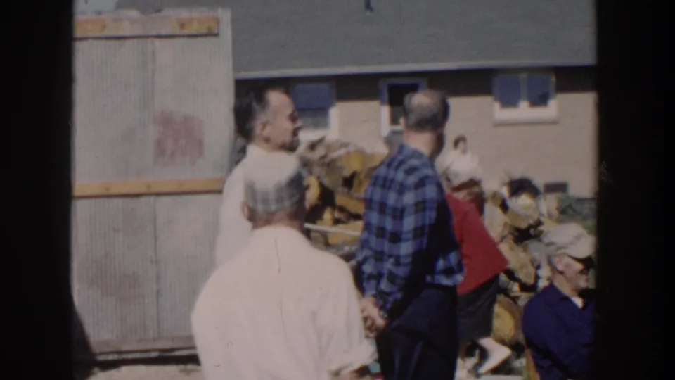 1958 Wisconsin frame showing adults gathered near a backyard woodpile and brick house, including a person in a red garment moving through the scene.