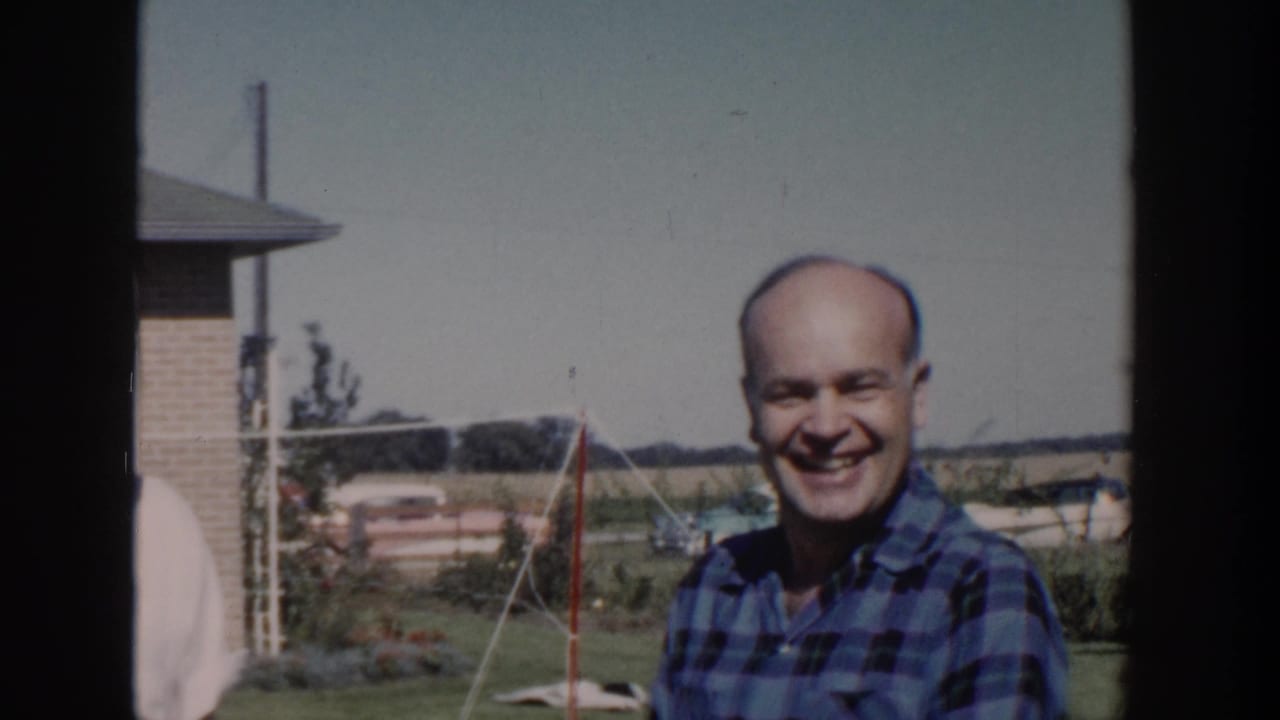 In 1958 Wisconsin home-movie footage, a smiling adult man in a blue plaid shirt stands in a suburban backyard.