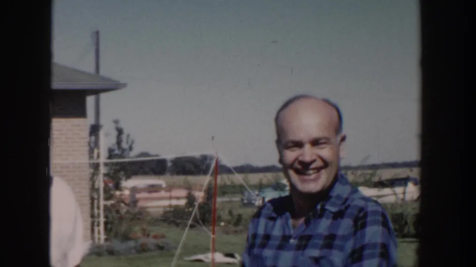 In 1958 Wisconsin home-movie footage, a smiling adult man in a blue plaid shirt stands in a suburban backyard.