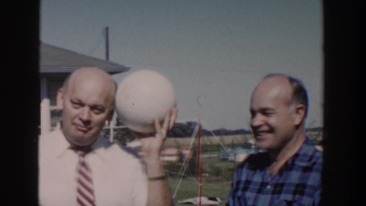 Two adult men stand in a Wisconsin backyard, with one in a tie lifting a large white ball beside his face.