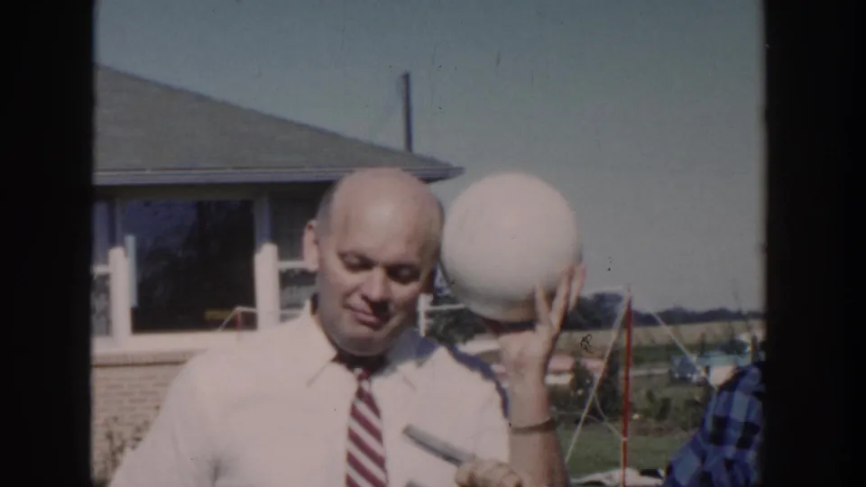 A man in a white shirt and striped tie holds a large white ball in a 1958 Wisconsin backyard scene.