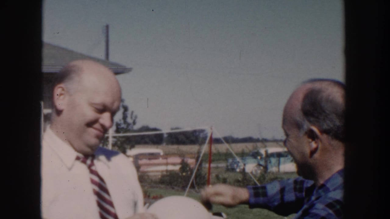 1958 Wisconsin home-movie frame of two bald adult men in a backyard, with one smiling in a white shirt and striped tie.