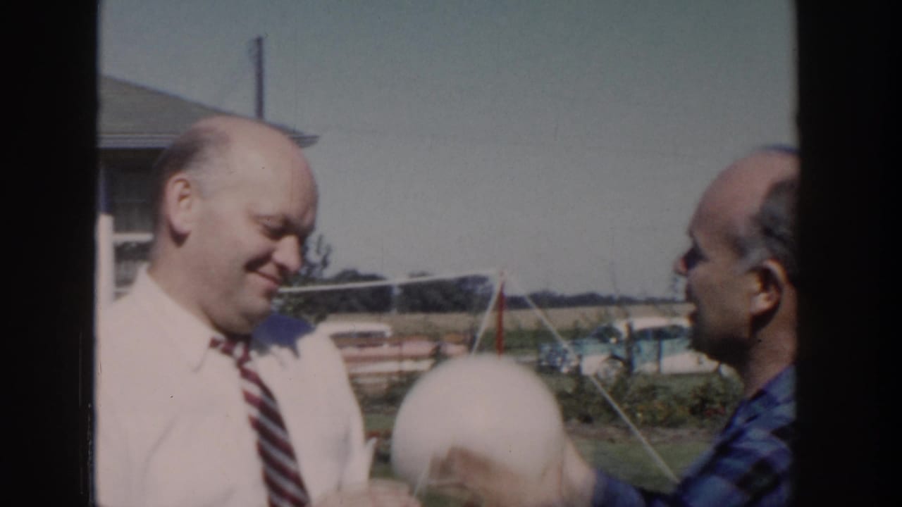 1958 Wisconsin backyard frame with two bald men, one smiling in a tie while the other raises or positions a pale round object near him.