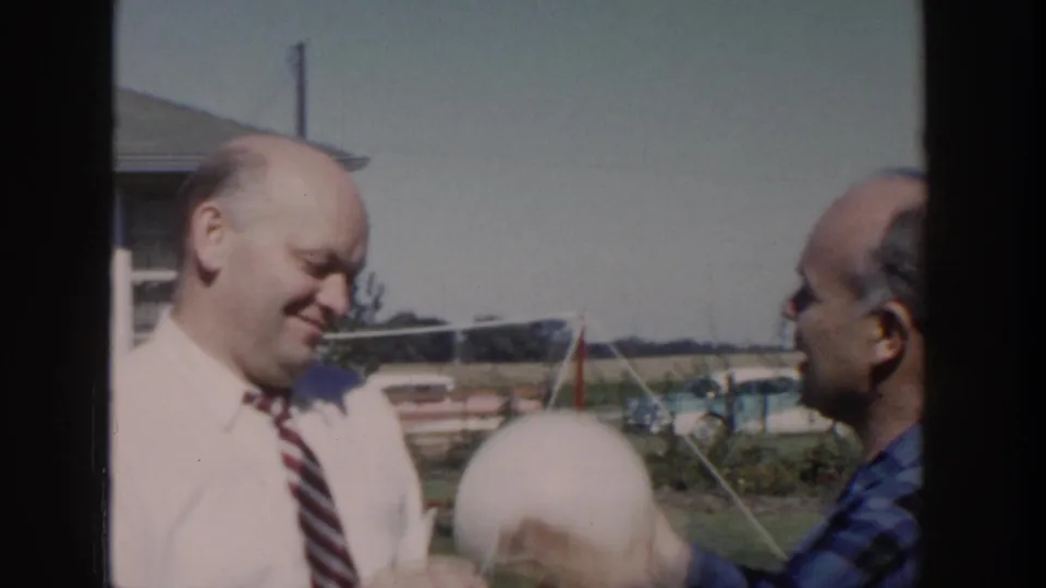 1958 Wisconsin backyard frame with two bald men, one smiling in a tie while the other raises or positions a pale round object near him.