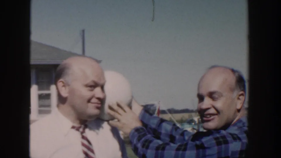 1958 Wisconsin home-movie frame of a smiling man in a blue plaid shirt holding a pale round object against another bald man's head.
